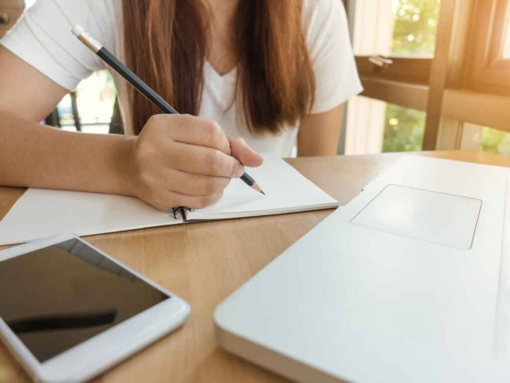 Garota escrevendo o esboço de uma redação em um caderno. Seu notebook esta ao seu lado, assim como seu celular.