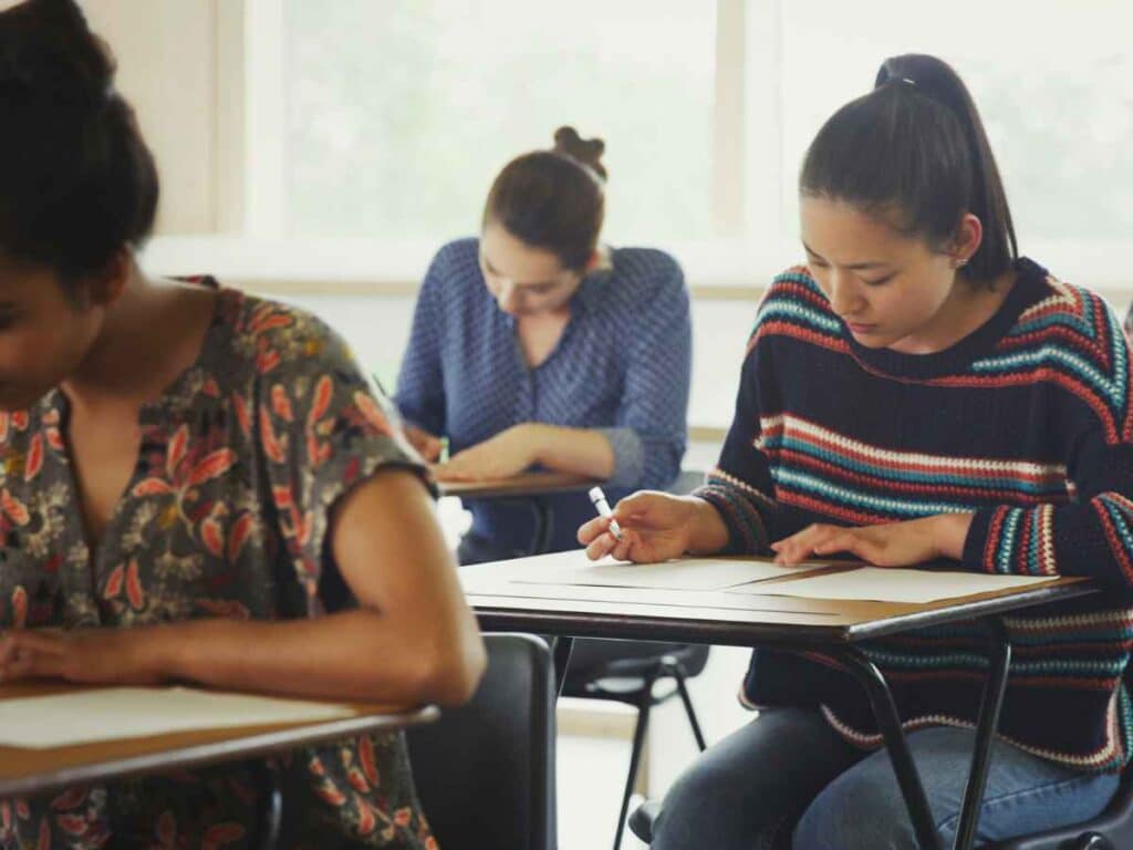 Estudantes concentradas fazendo uma prova em sala de aula. Sentadas em carteiras escolares, elas escrevem em folhas de papel sob clima silencioso e atento, típico de momentos avaliativos.