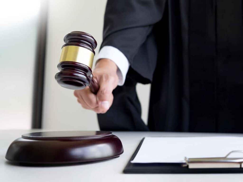 Juiz em toga preta segura martelo de audiência prestes a bater sobre a base de madeira em um tribunal.