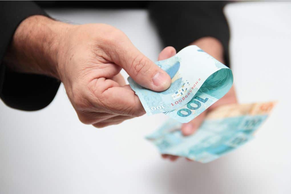 Mão de home entregando notas de 100 reais para alguém, como se estivesse comprando algo.