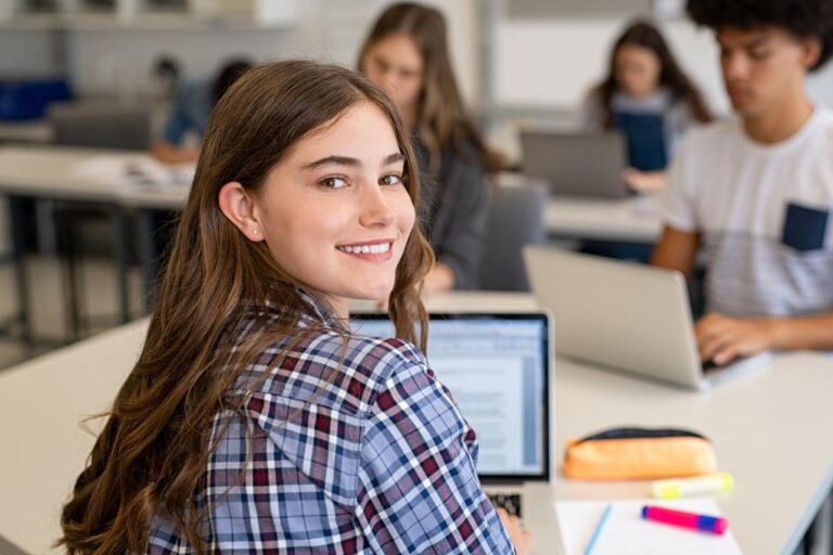 Jovem sorridente em sala de aula com notebook, ao lado de outros adolescentes estudando.