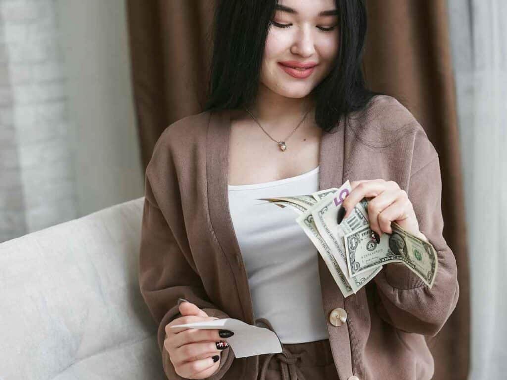 Mulher sentada segurando cédulas com expressão de satisfação ao observar uma nota específica, como se tivesse acabado de encontrar um exemplar raro para compor sua coleção.