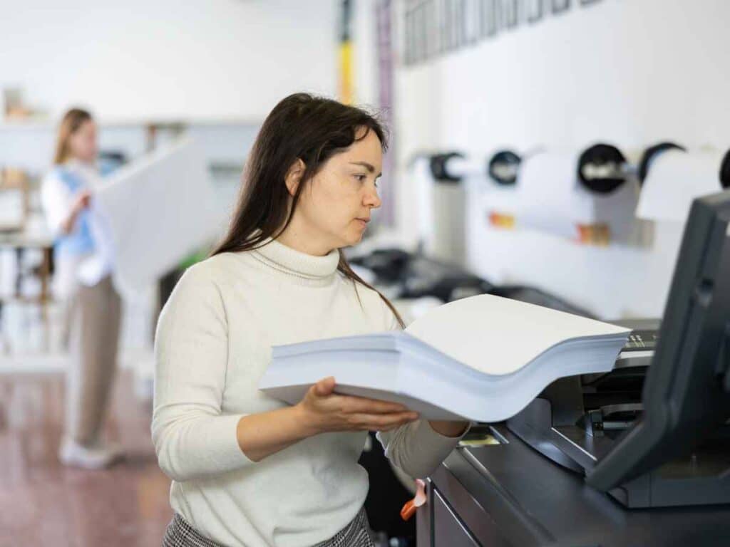 Mulher colocando uma grande resma de papel em uma impressora em ambiente de escritório ou gráfica. Ela está concentrada na tarefa, usando suéter branco e cercada por equipamentos de impressão. Ao fundo, outra pessoa lida com folhas de papel, reforçando o cenário profissional.