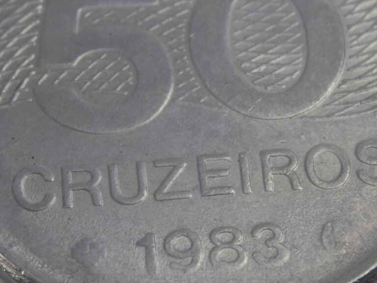 Imagem em close-up de uma moeda brasileira de 50 cruzeiros do ano de 1983. É possível ver em destaque os números “50”, a palavra “CRUZEIROS” e o ano de emissão gravados na superfície metálica da moeda, com textura detalhada.