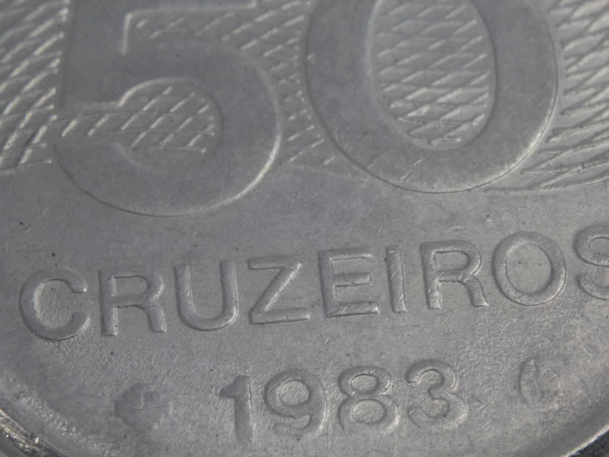 Imagem em close-up de uma moeda brasileira de 50 cruzeiros do ano de 1983. É possível ver em destaque os números “50”, a palavra “CRUZEIROS” e o ano de emissão gravados na superfície metálica da moeda, com textura detalhada.