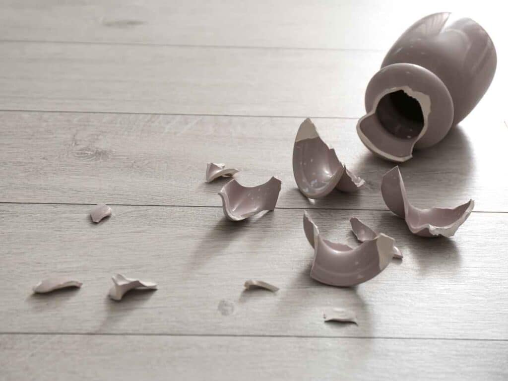 Vaso de cerâmica quebrado em vários pedaços sobre um piso claro de madeira. 