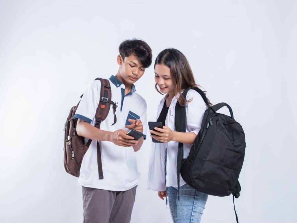 Dois adolescentes com mochilas interagindo com seus celulares, em frente a um fundo branco.
