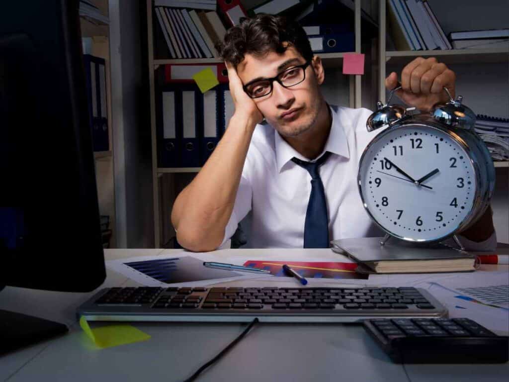 Homem com expressão cansada segurando um grande relógio, representando a exaustão por longas jornadas de trabalho em escritório.