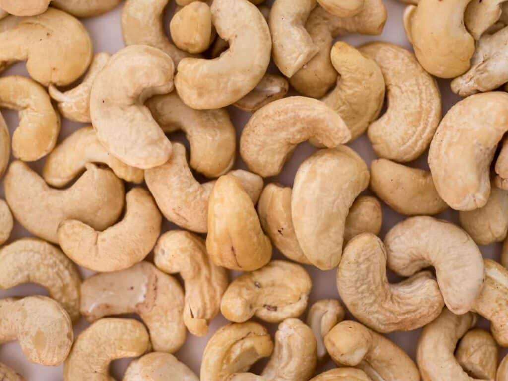 Vista aproximada de várias castanhas de caju torradas, com coloração bege clara e textura levemente enrugada, espalhadas sobre uma superfície branca.