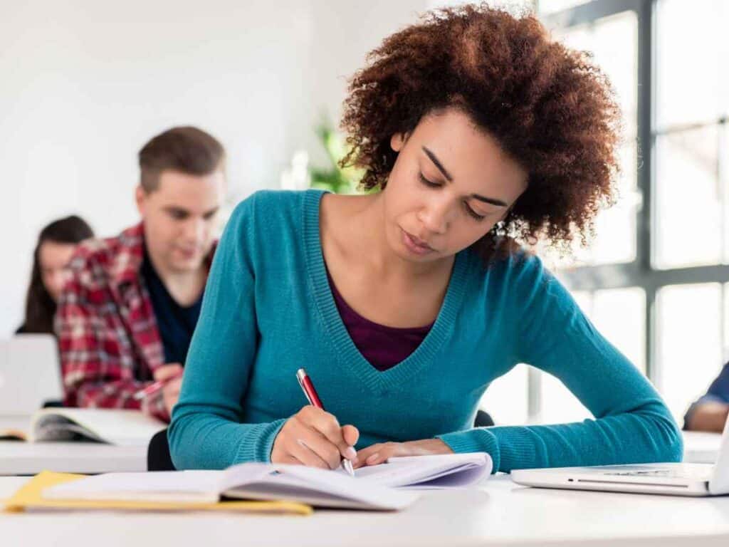 Jovem concentrada escrevendo em um caderno durante aula, com livros e notebook sobre a mesa. Cena remete à preparação para a redação do Enem 2025.