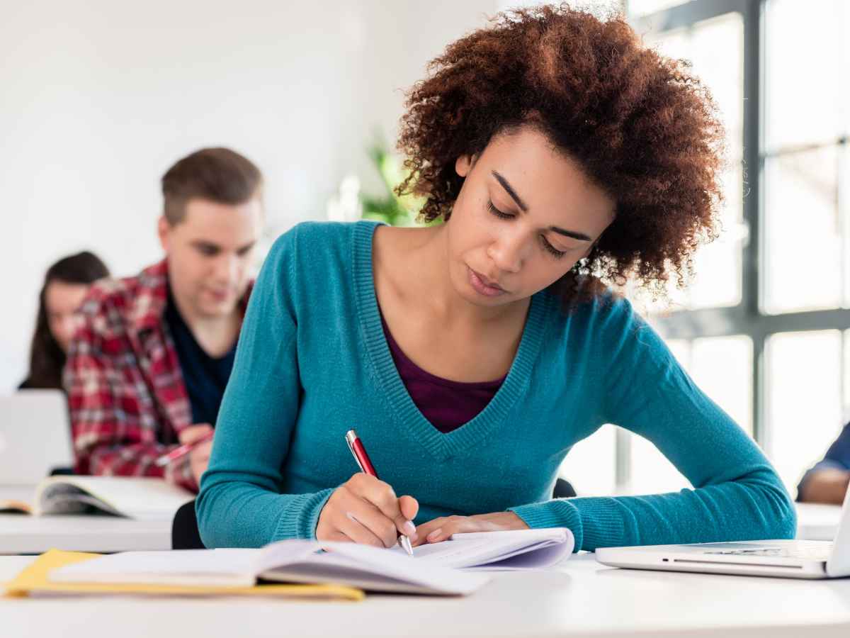 Jovem concentrada escrevendo em um caderno durante aula, com livros e notebook sobre a mesa. Cena remete à preparação para a redação do Enem 2025.