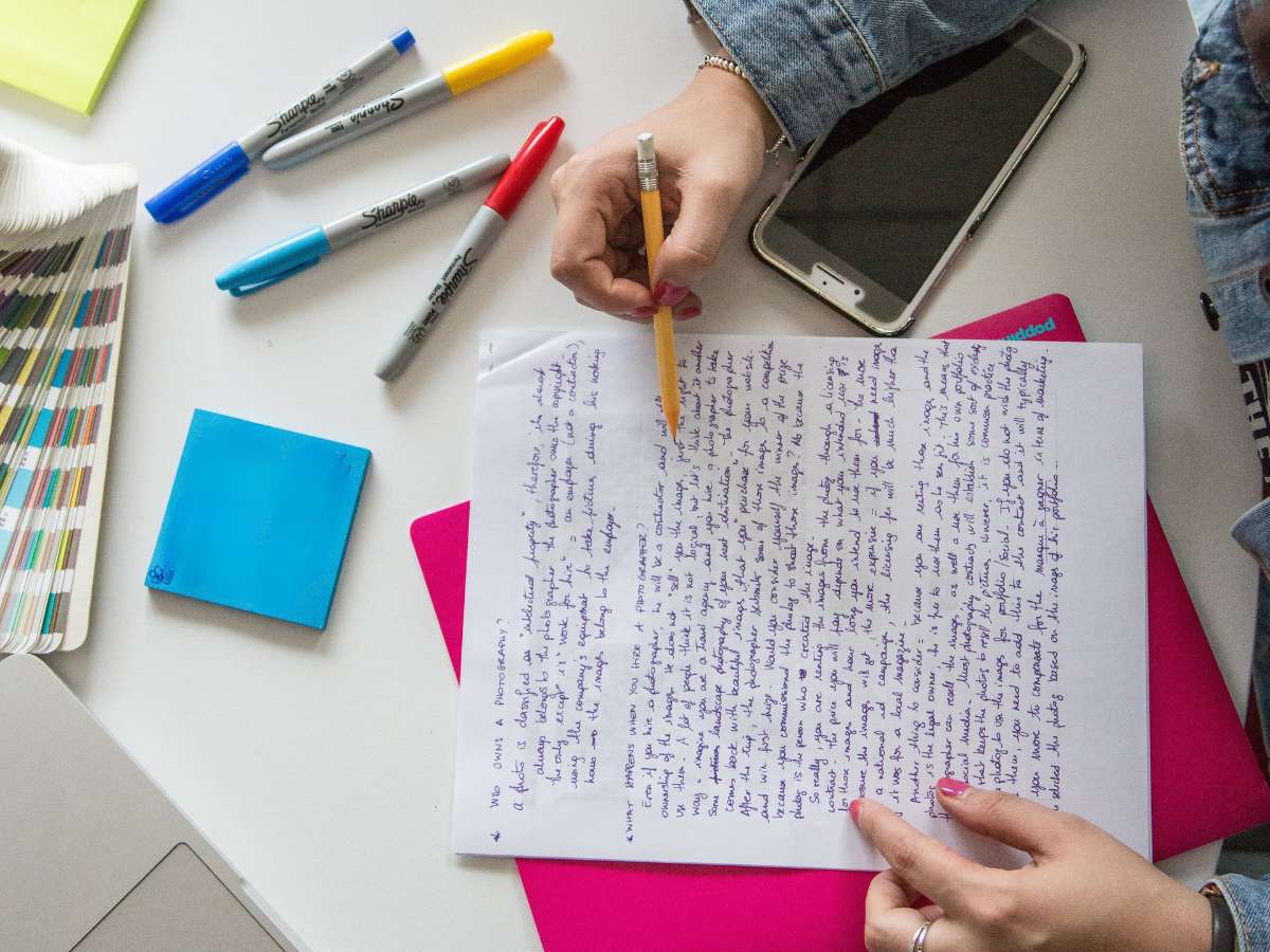 Mão feminina segura lápis sobre folha de papel com texto manuscrito, em mesa com marcadores coloridos, celular, óculos e caderno rosa ao fundo, representando atividade de interpretação de texto.