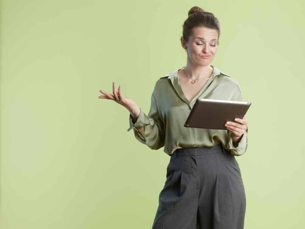 Mulher em pé, segurando um tablet com expressão de dúvida enquanto lê, em frente a um fundo verde claro.