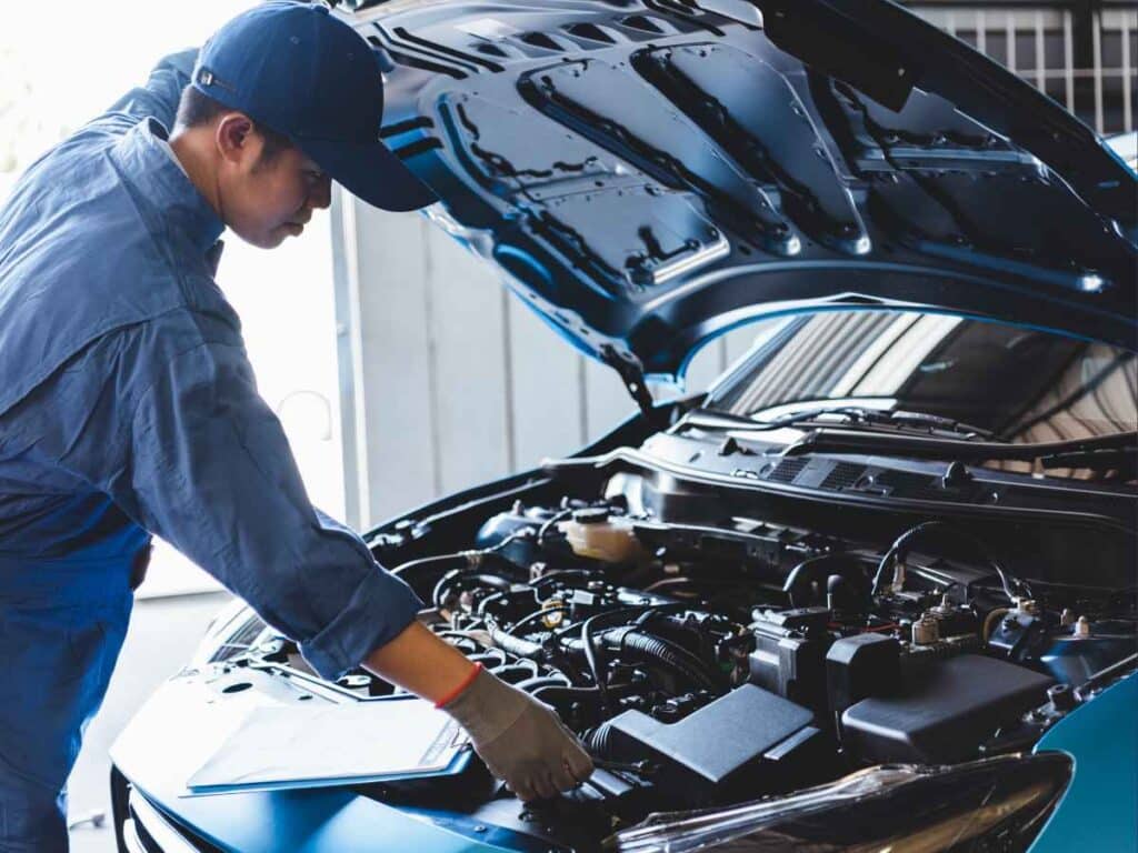 Mecânico usando uniforme azul e boné realiza inspeção no motor de um carro com o capô aberto em uma oficina.